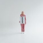A woman in a pink suit holds a 'Women' sign, symbolizing empowerment in a minimalist studio.